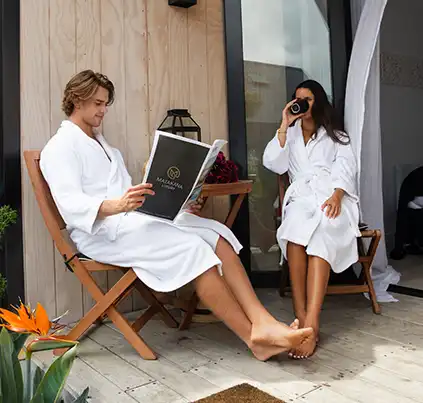 A couple relaxing in robes on the outdoor deck at Matakana Luxury, enjoying coffee and reading outside their villa.